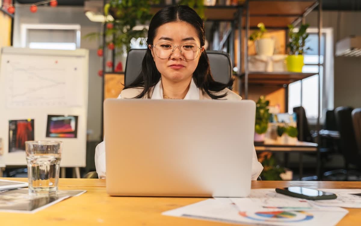 Một người đeo kính ngồi làm việc với laptop, phía sau là bảng biểu đồ và kệ cây xanh