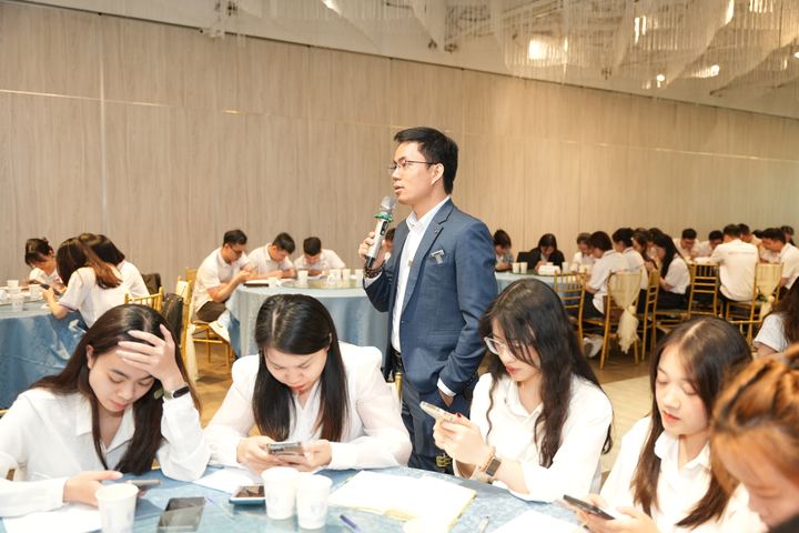 Diễn giả cầm micro giữa hội trường, người tham dự ghi chép