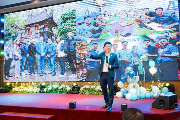 Diễn giả trên sân khấu, phía sau là màn hình ảnh tập thể